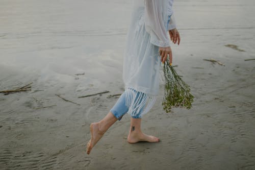 Person Standing Barefoot on Beach while Holding Flowers