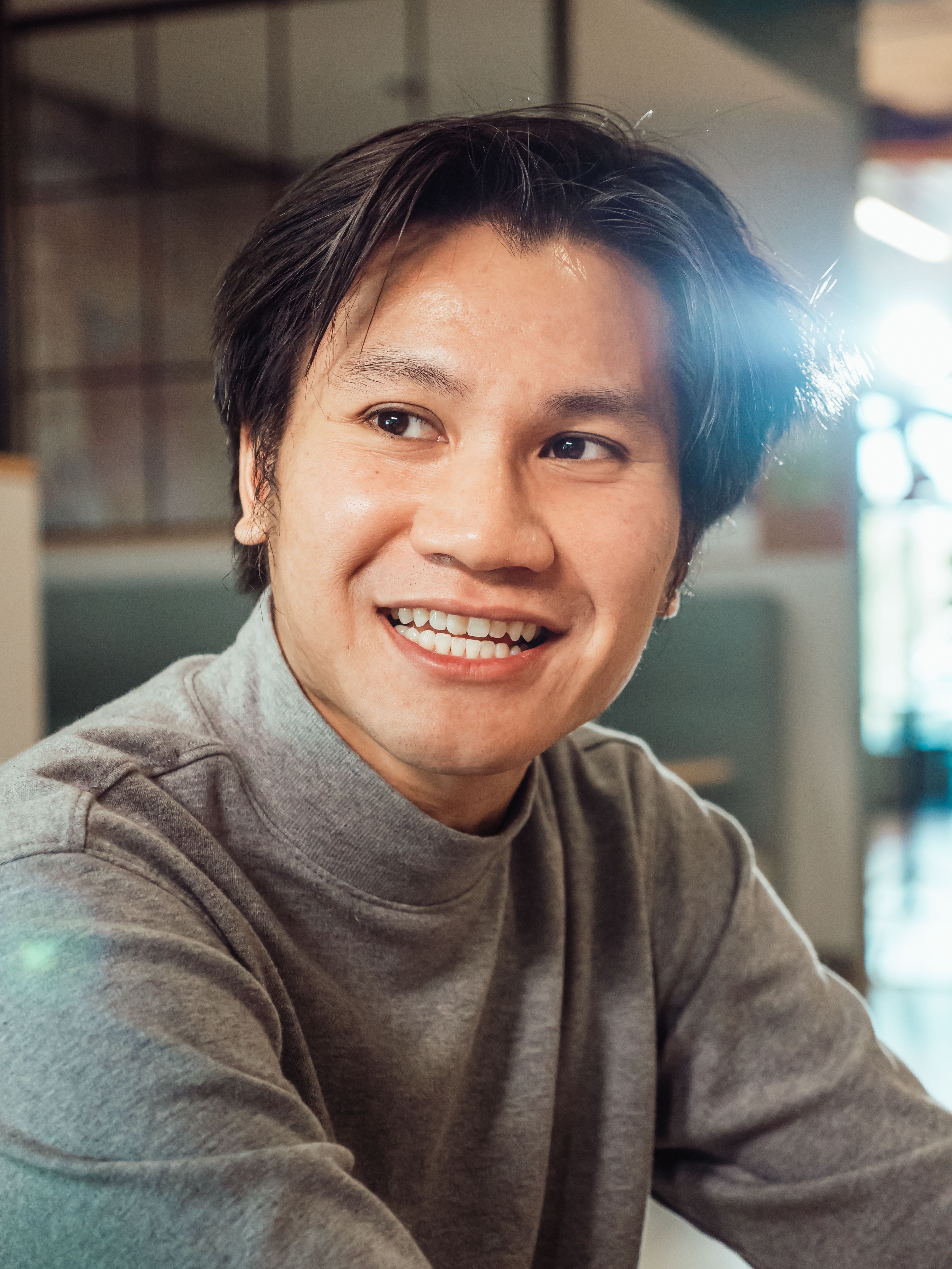 Asian man in a sweater smiling warmly indoors, captured in natural light.