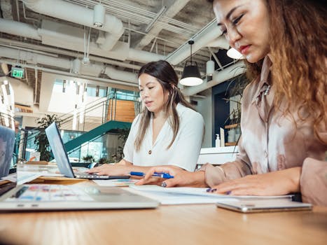 Two women working together on laptops and documents in a bright, modern office setting.