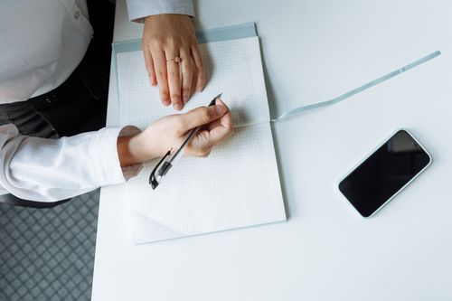 Free Close-up of hands writing in a notebook with a smartphone on a white desk. Stock Photo