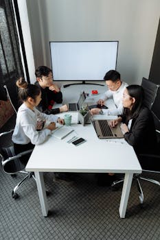 A group of professionals discussing a project in a modern office setting with laptops and a whiteboard.