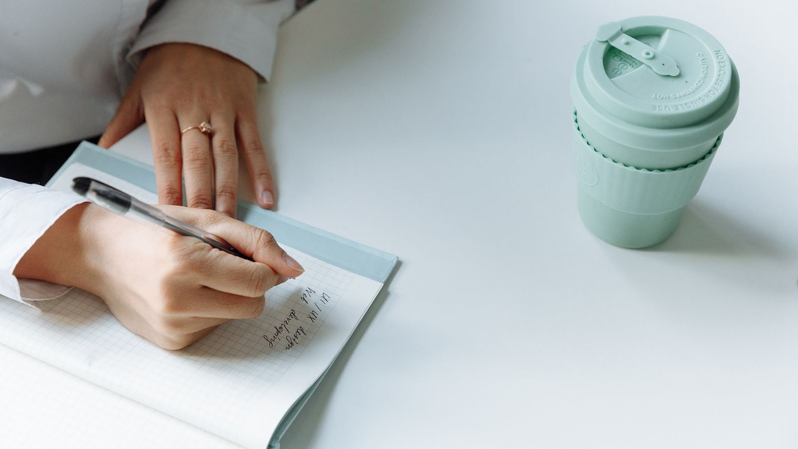 Hands writing in a squared notebook beside a green reusable cup