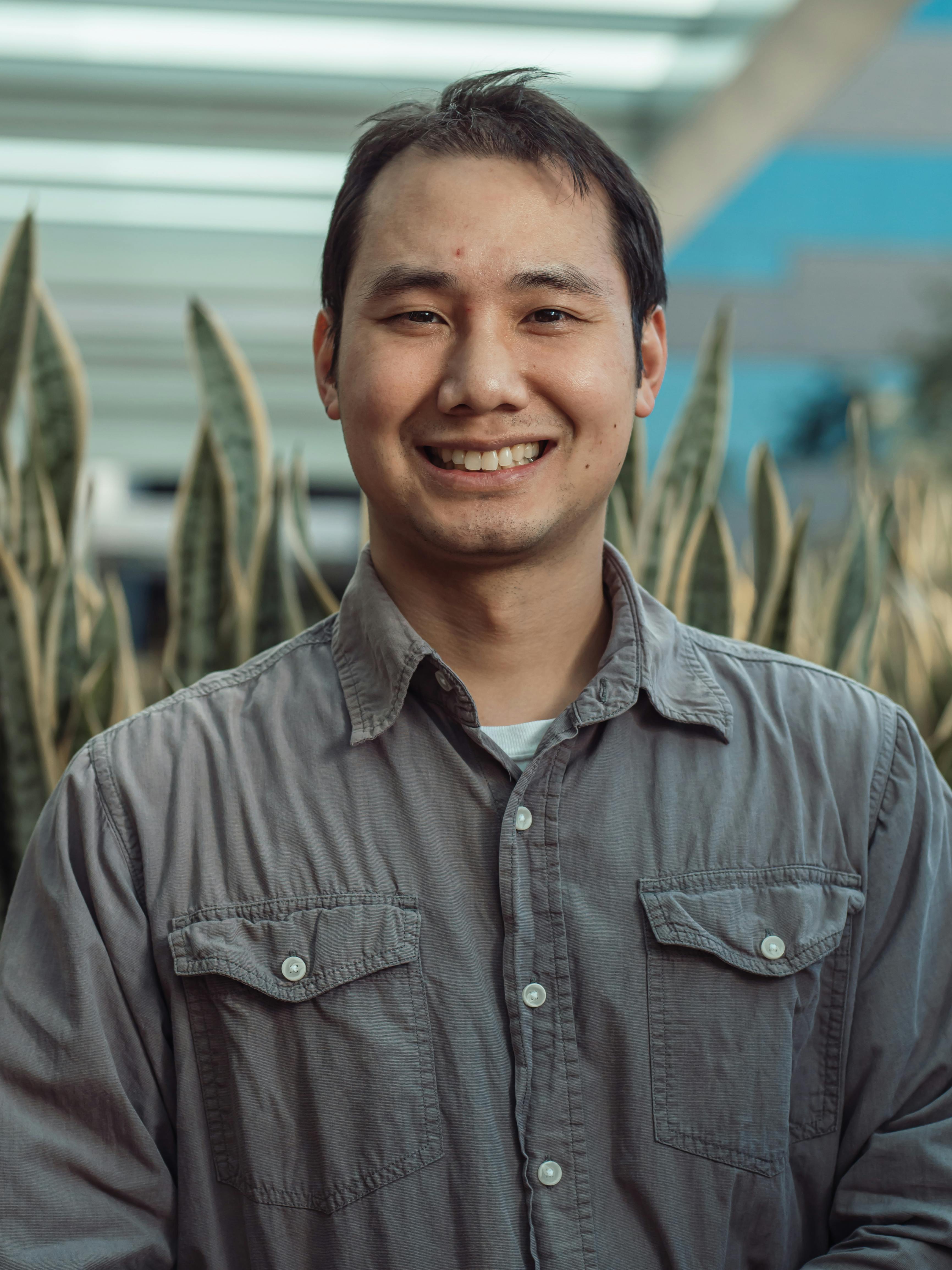 A smiling adult male in a casual setting, captured indoors with natural plants in the background.