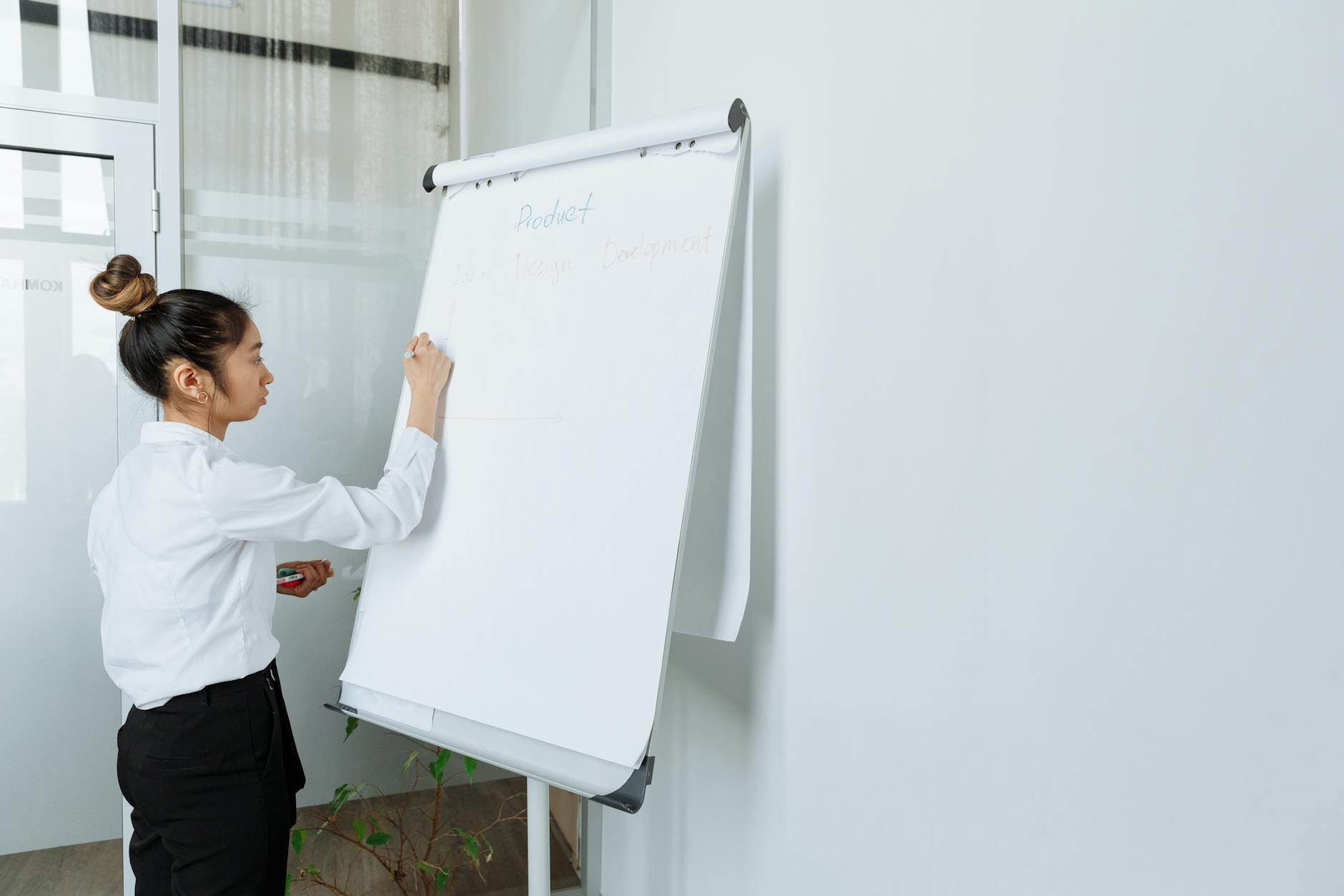 Businesswoman in white blouse writing on a flipchart during a presentation in a modern office setting.