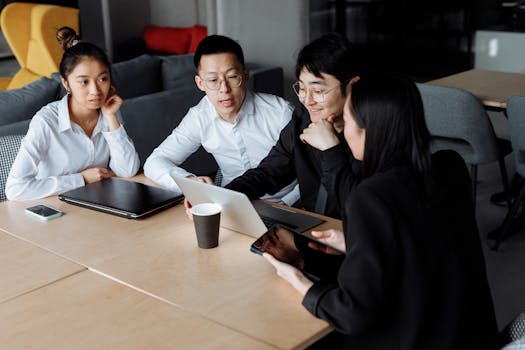 A group of colleagues engaged in a business meeting discussing plans around a laptop in a modern office.