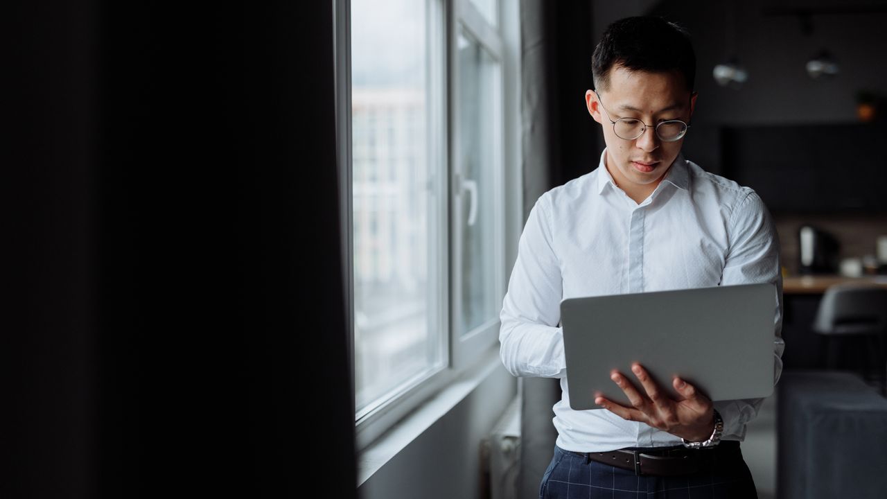 An Asian man in a white shirt using a laptop while standing by a window indoors.