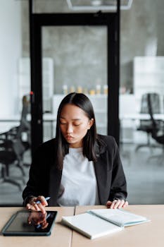 Asian businesswoman using digital tablet and notebook in a modern office setting, focused on planning tasks.