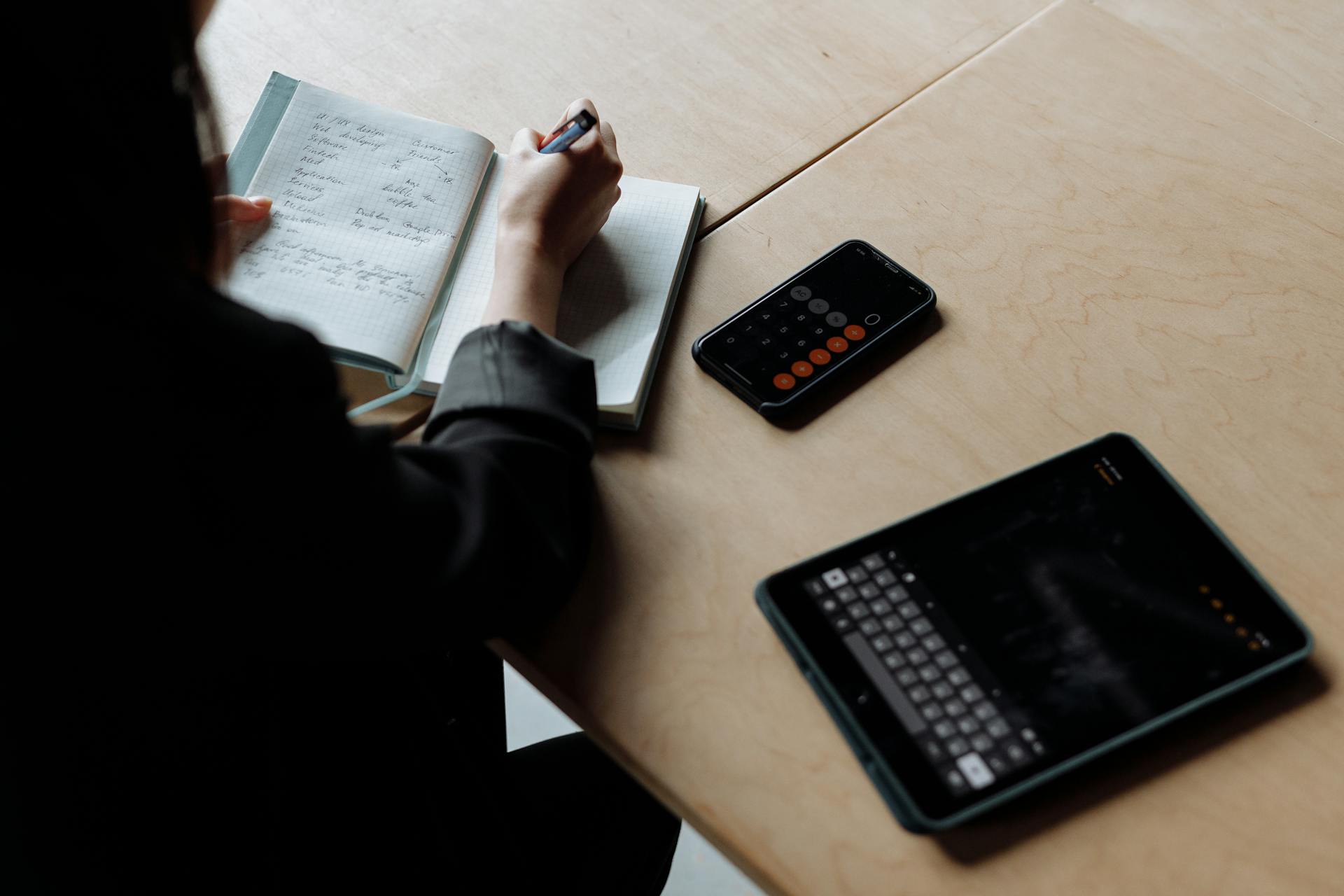 Person in office setting writing in notebook with tablet and smartphone on desk, emphasizing modern work.