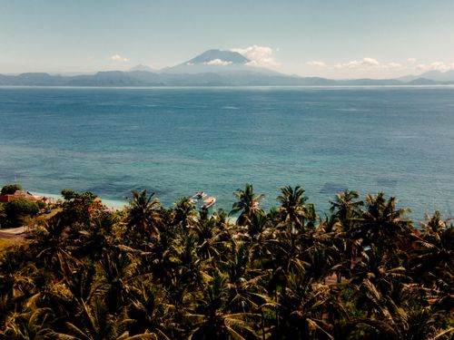 Free A stunning aerial view of the Indonesian coastline featuring lush palm trees and vibrant ocean waters with distant mountains. Stock Photo