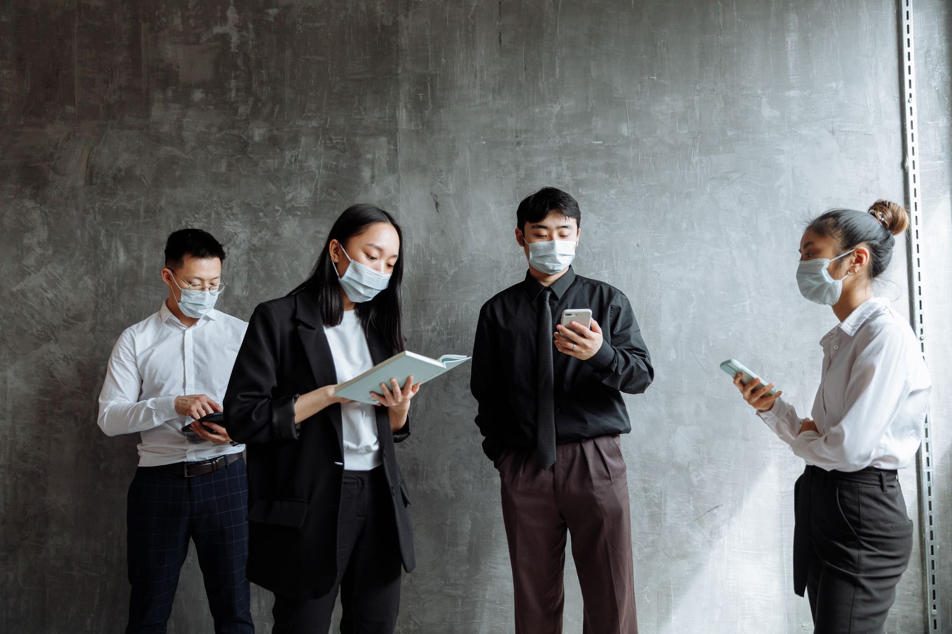 Group of professionals wearing masks in an office setting, using mobile devices and notebook.