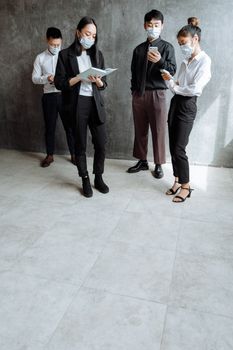 A group of Asian professionals wears face masks while reading books and smartphones indoors.