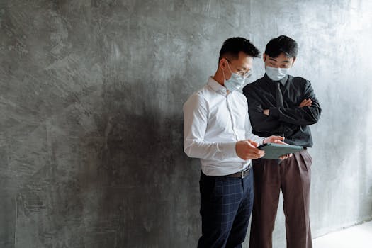 Two men in face masks using a tablet, discussing business indoors.