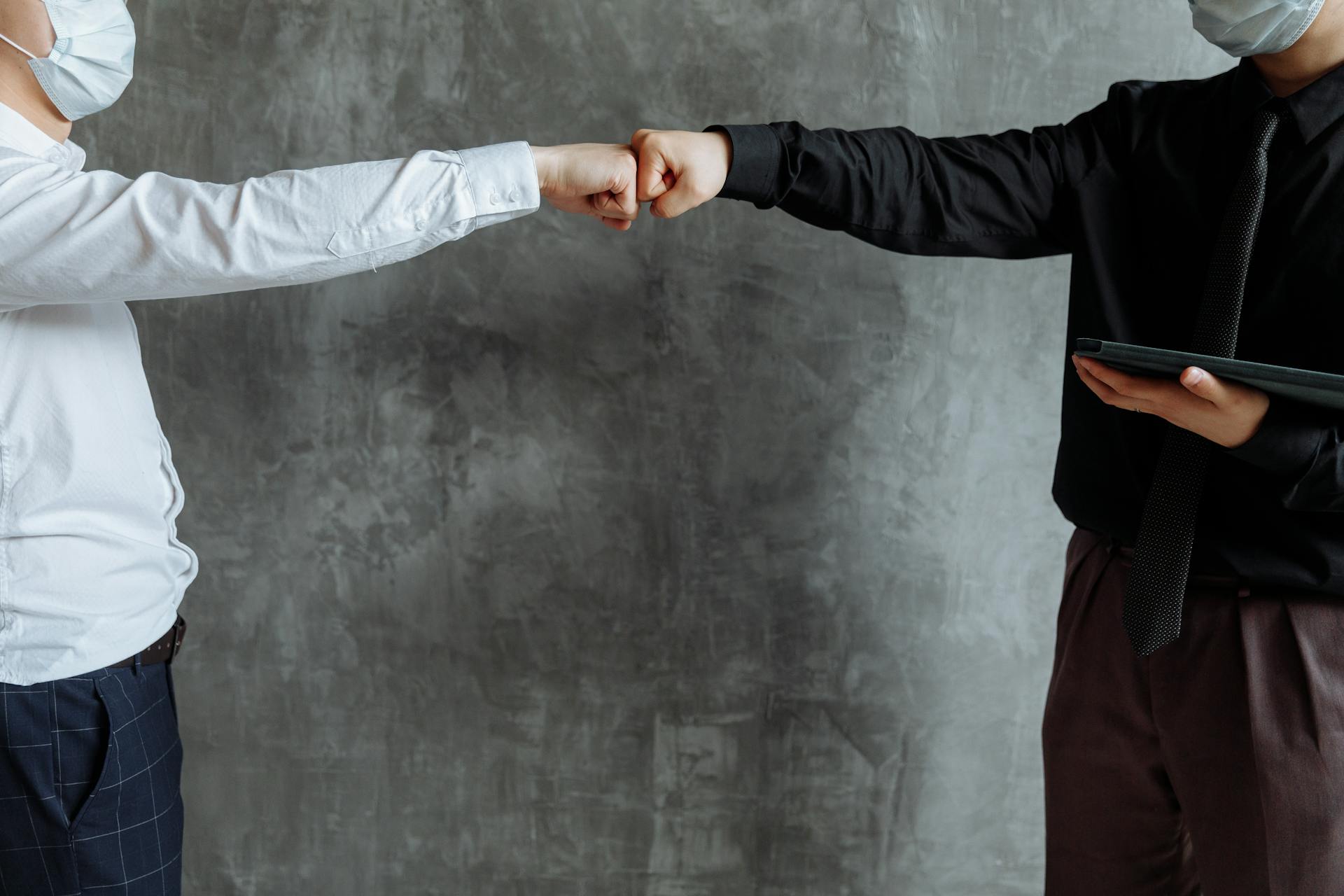 Two business professionals in masks fist bumping indoors, symbolizing cooperation and safety.