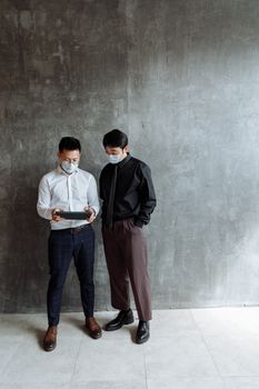 Two men in business attire and face masks using a tablet indoors.