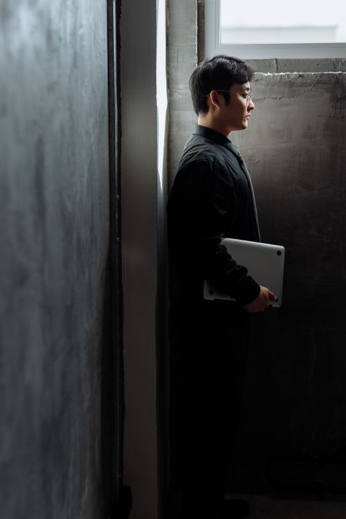 Free Asian man in profile holding laptop, standing in a dimly lit concrete room with a window. Stock Photo