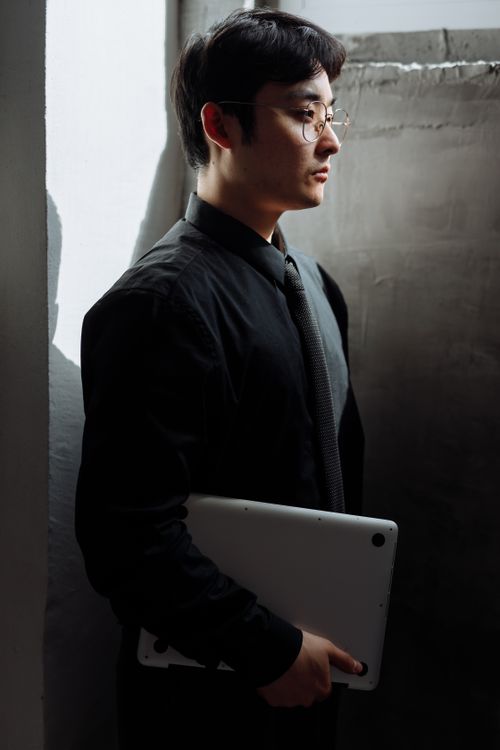 Free Asian man in black suit holding a laptop, standing by a concrete wall. Professional and contemplative mood. Stock Photo