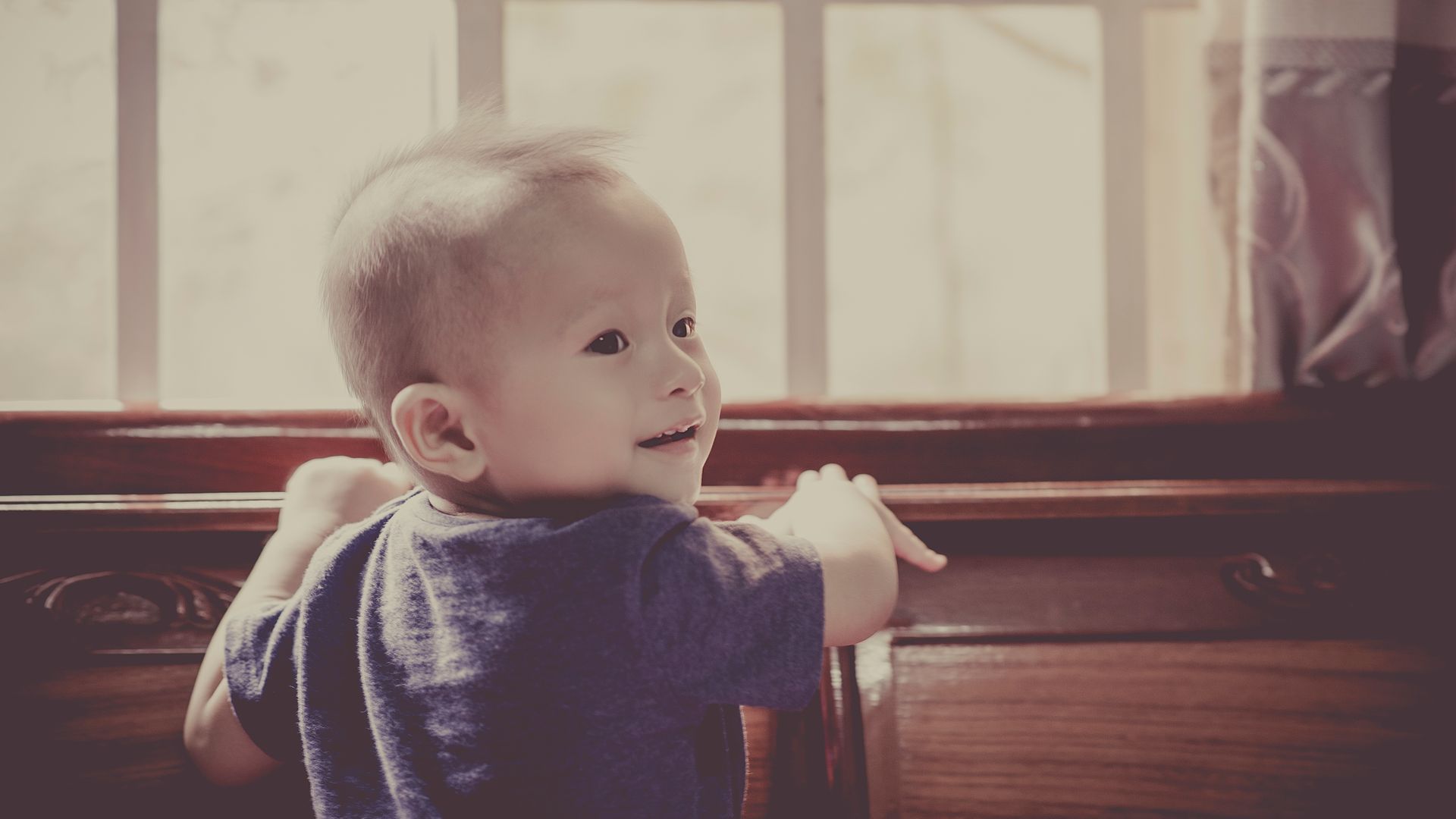 Charming baby smiling joyfully while leaning on wooden furniture by the window.