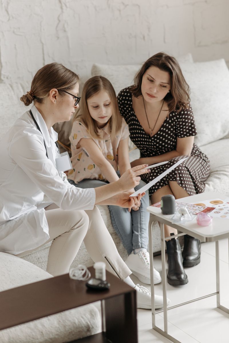 Médica mostra exames para mãe e filha durante consulta em ambiente acolhedor, sentadas no sofá do consultório