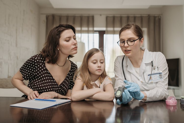 Doctor Sitting At The Table With A Mother And Daughter And Holding A Glucometer 