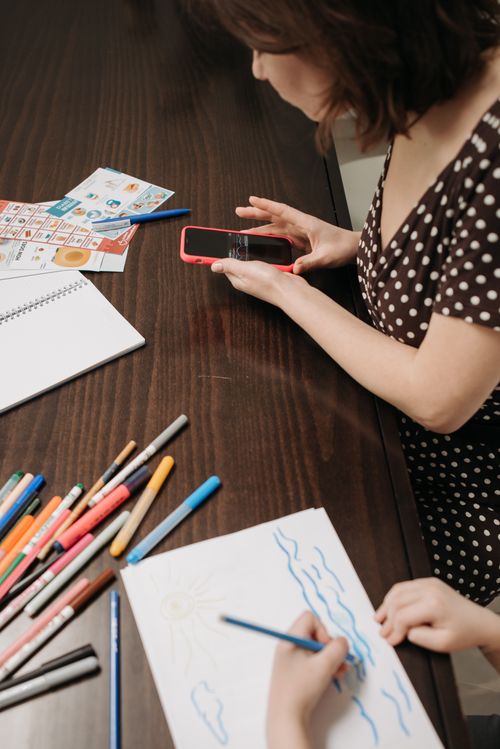 Free A mother supervises her child drawing at a table while holding a smartphone in a cozy indoor setting. Stock Photo