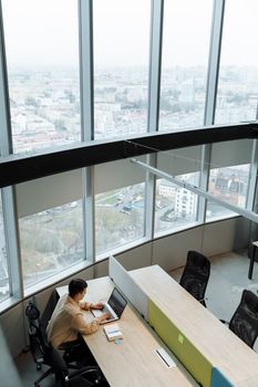 A man working on a laptop at a modern office desk with panoramic city views through large windows.