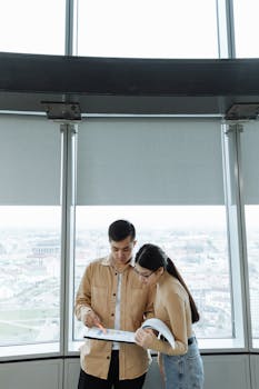 Two colleagues reviewing documents near a large window in an office setting.