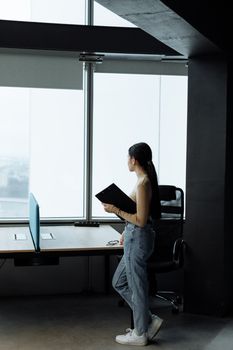 Young woman in denim pants holding a binder, looking out office window.
