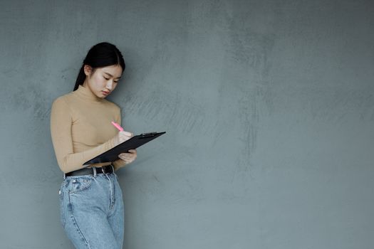 Asian woman writing notes on a clipboard standing against a minimalist gray wall.