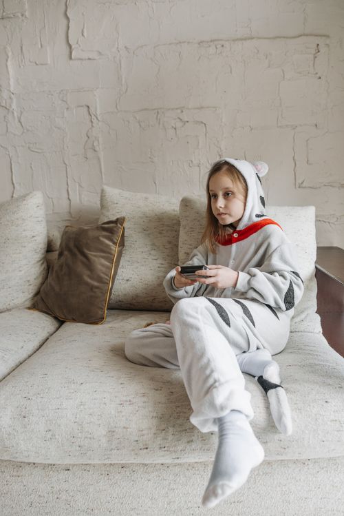 Free Young girl in cute pajamas sitting on a sofa, focused on playing a video game indoor. Stock Photo