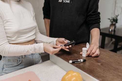 Close-up Shot of a Woman in Beige Long Sleeves Preparing the Glucose Meter while Standing Beside a Person Wearing Black Hoodieb