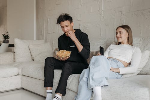 A Woman Sitting on a Couch while Looking at the Man Eating Chips