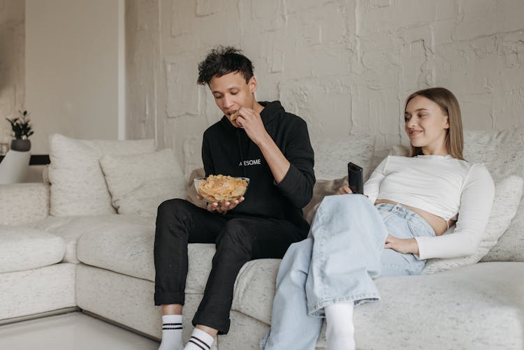 A Woman Sitting On A Couch While Looking At The Man Eating Chips