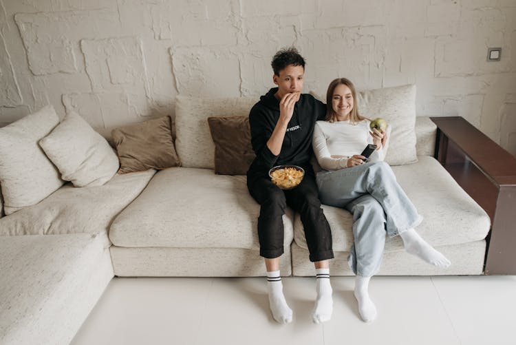 A Couple Sitting On A Couch While Eating Snacks