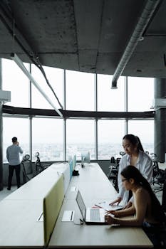 Asian women collaborate on a project in a modern office setting, utilizing laptops for teamwork.