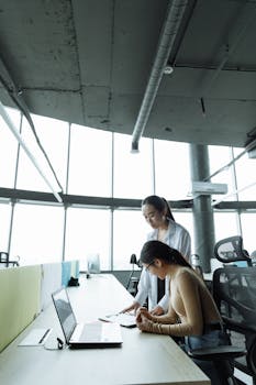 Two women collaborate at a desk in a modern office with large windows.