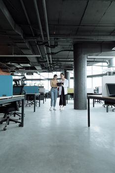 Two women discussing work in a modern open-plan office, showcasing teamwork and collaboration.