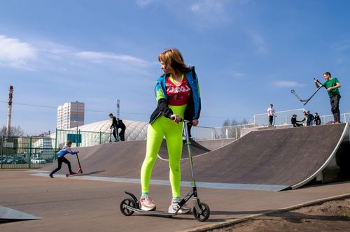 Free Full body of girl in sportswear standing in skatepark with kick scooter and looking away while other teenagers riding on background in sunny day Stock Photo