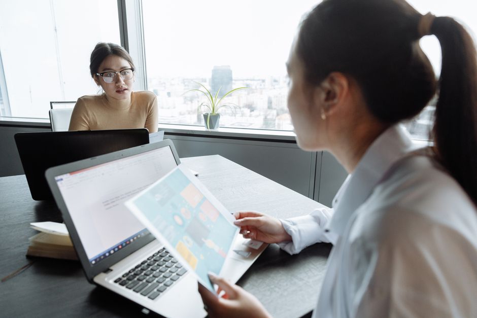 Two businesswomen discussing project ideas in a modern office setting with laptops.