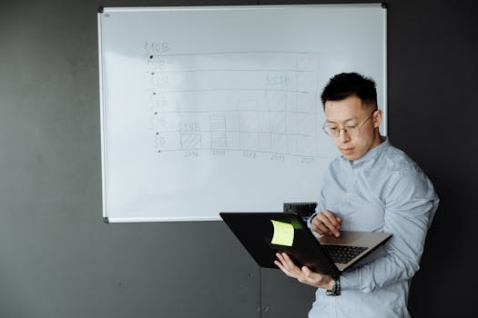 An Asian man reviewing business data on a laptop beside a whiteboard in a modern office setting.