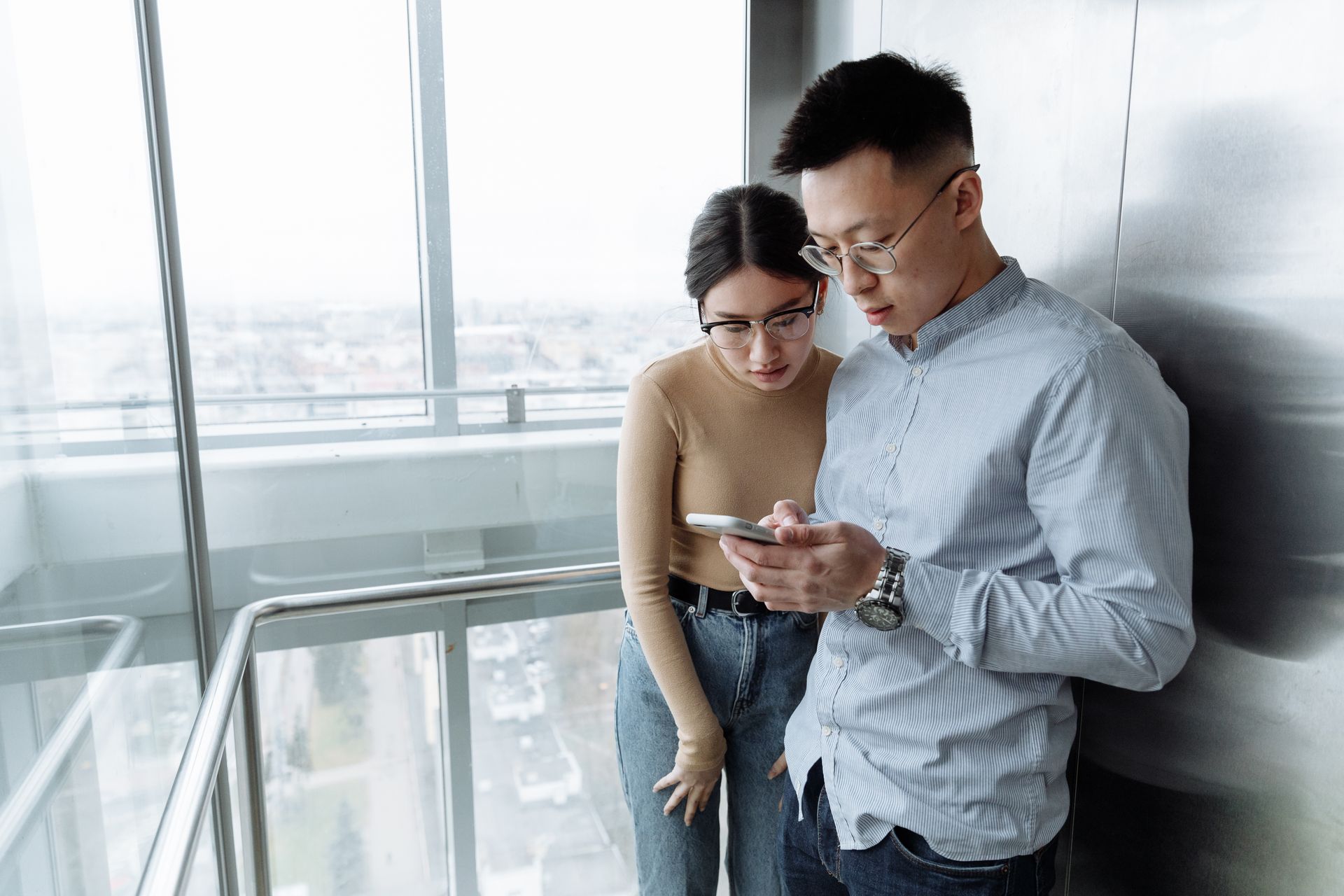 Asian man and woman looking at smartphone in modern glass elevator with city view.