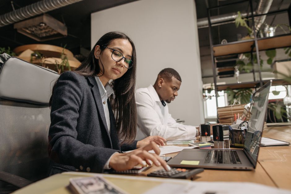A busy office scene with two professionals working on laptops and calculators.