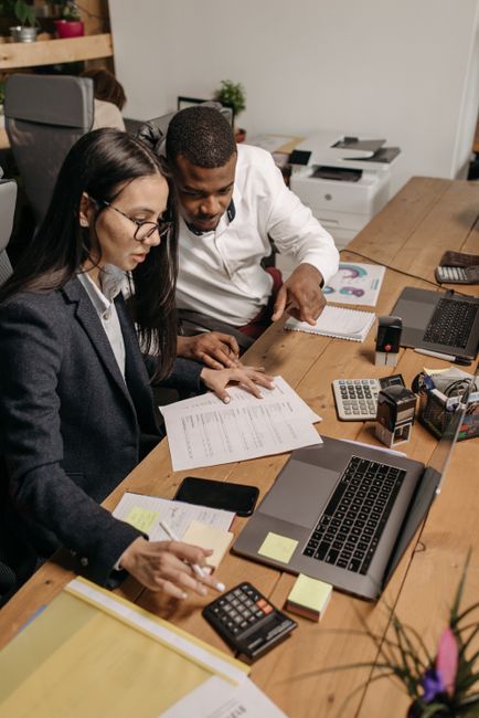 Two employees working together on financial documents with laptops and calculators in an office setting. Two employees working together on financial documents with laptops and calculators in an office setting.