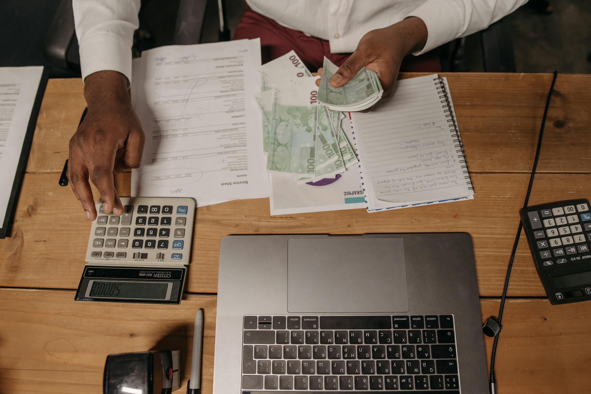 Man Sitting at the Desk and Counting Money while Using a Calculator