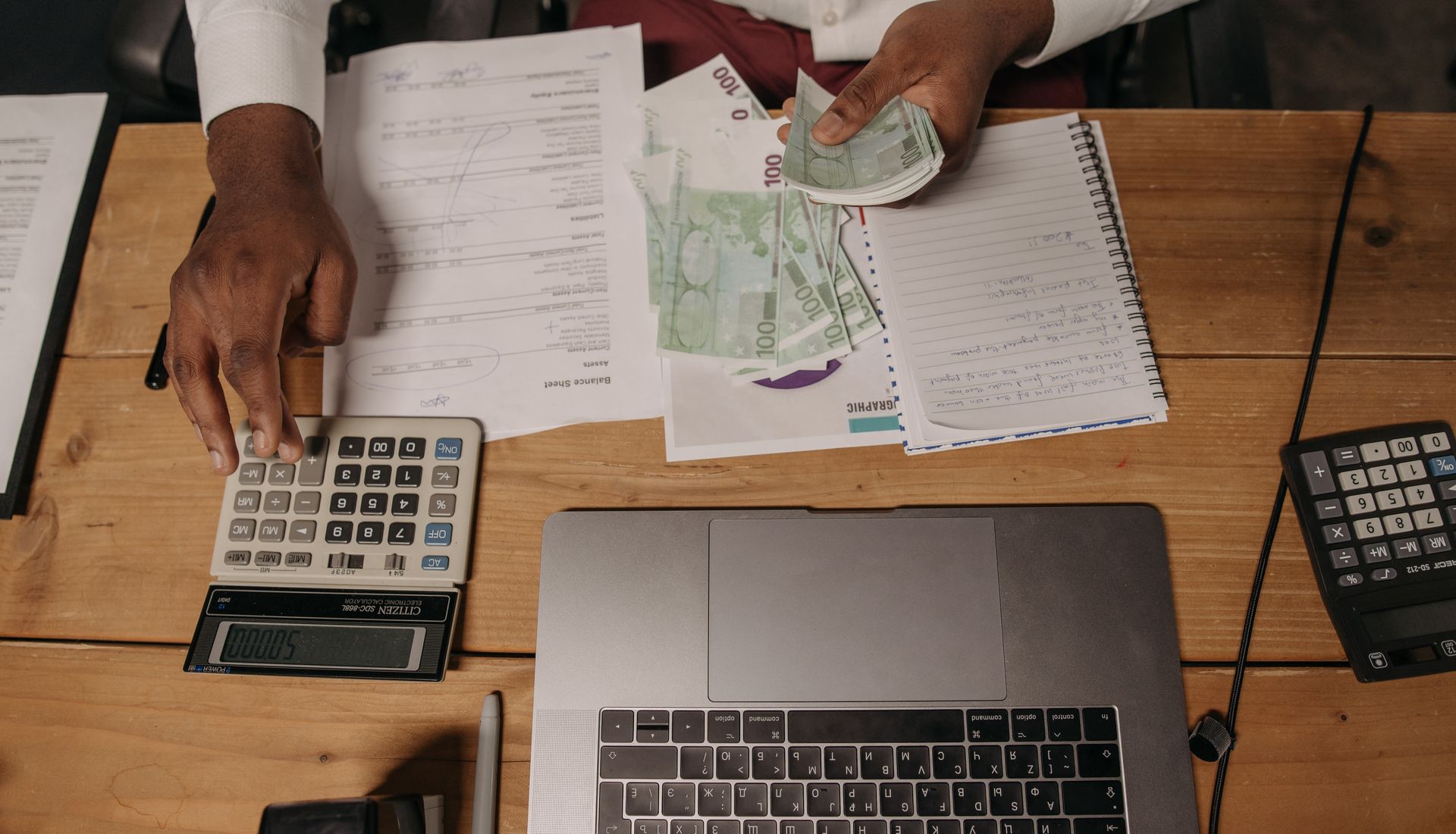 A person counting cash beside documents and a laptop