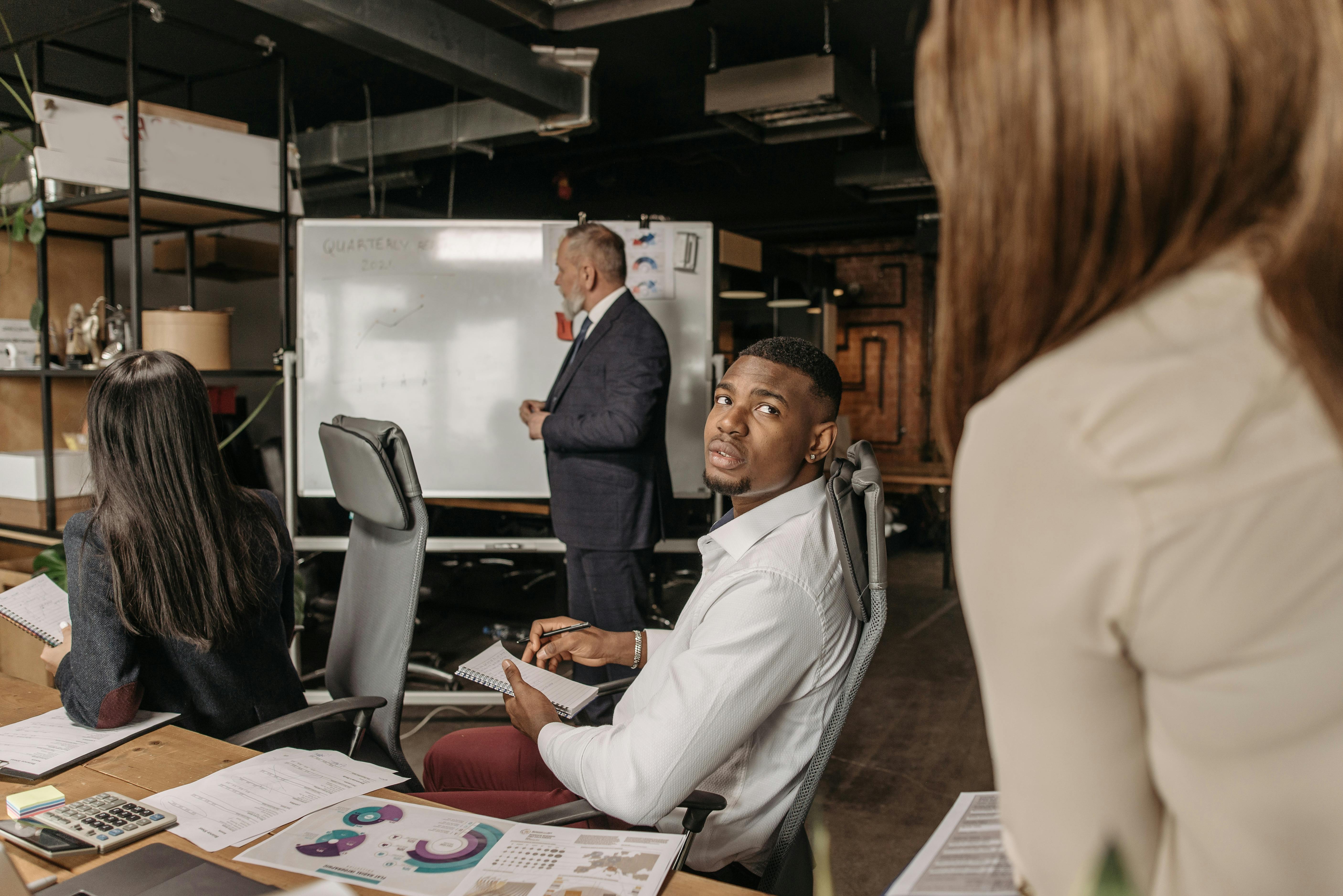 A diverse team collaborates in a modern office setting during a meeting with documents and a whiteboard.