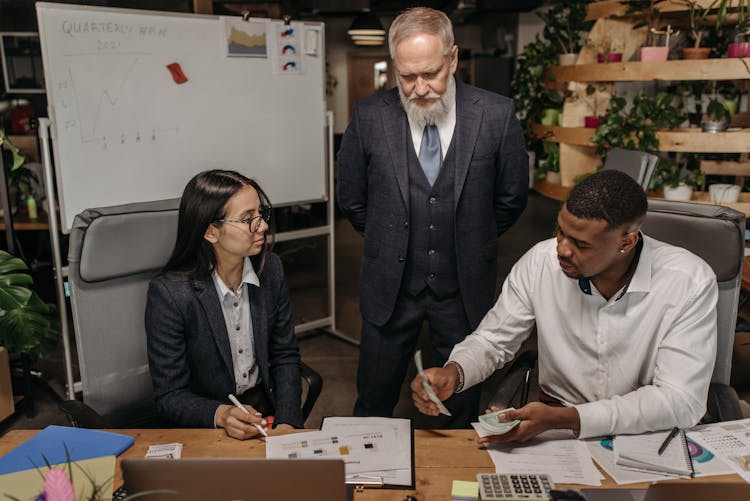 Boss Looking At Young Man And Woman Sitting At The Desk In An Office And Working 