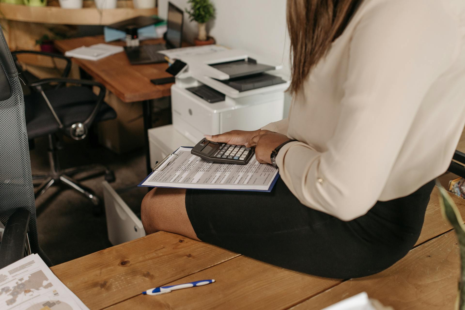 Professional woman sits at desk calculating finances with clipboard and calculator.