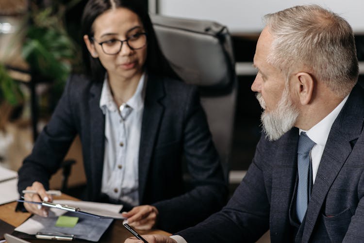 Elderly Man And Young Woman Sitting At The Desk In An Office And Talking 