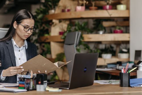 Free Businesswoman in office reviewing documents with a laptop, surrounded by plants and office supplies. Stock Photo