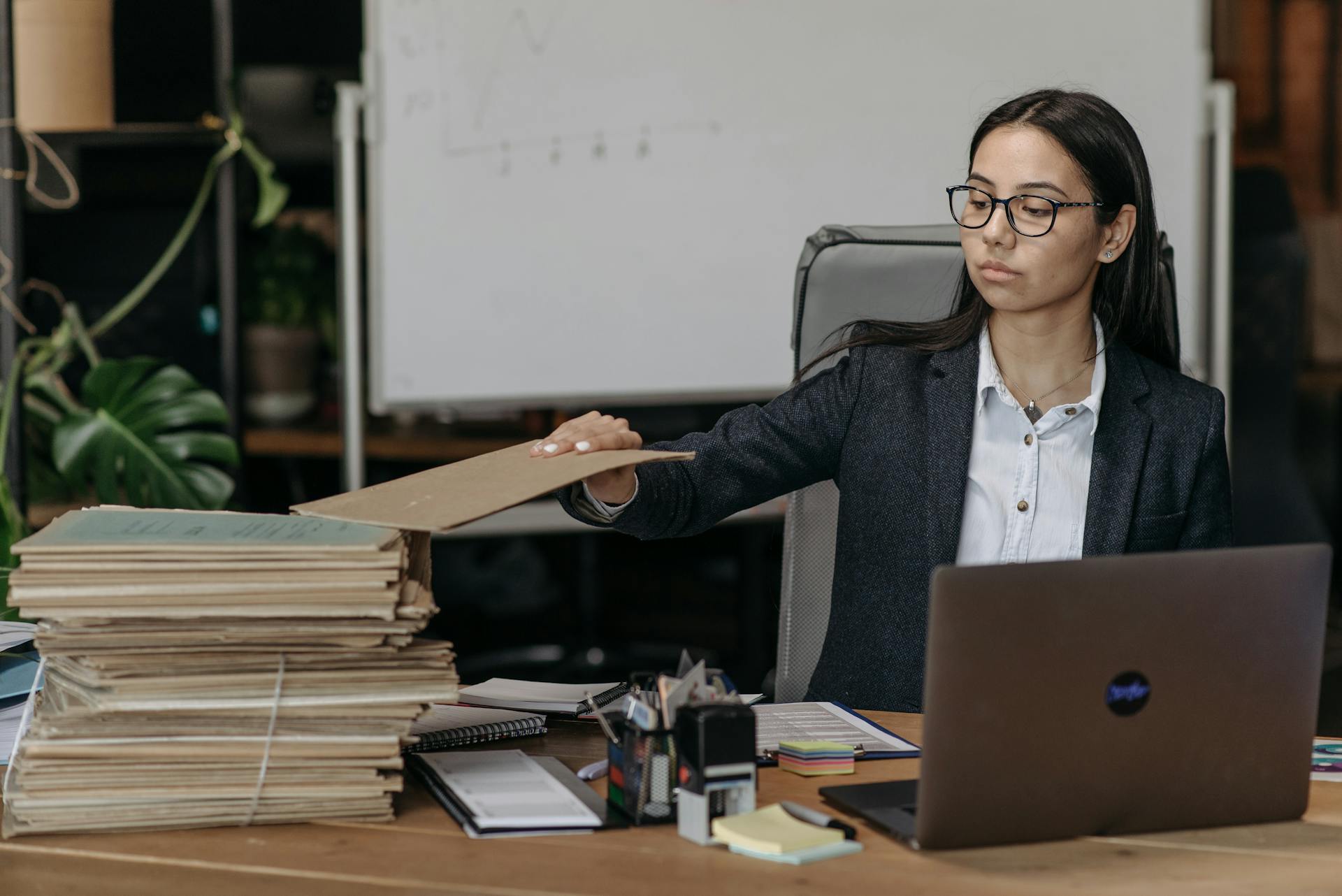 A businesswoman in a modern office organizing a stack of files beside a laptop.